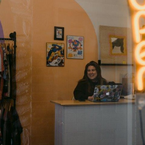 Woman smiles at cash register with laptop  in front and a glowing open sign to the right and full rack of clothes to the left