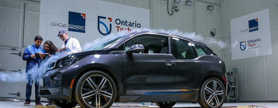 2 students and an instructor stand next to a vehicle looking at a clipboard one of the students is holding, with a black vehicle to their left that has a wind stream passing over the front of the vehicle. The ACE and Ontario Tech logos are on a white wall behind them. 