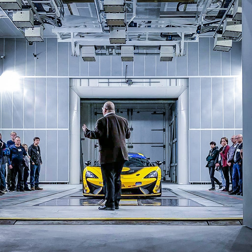 Man stands brightly lit in front of a yellow sports car, speaking to a crowd of individuals to the right and left.