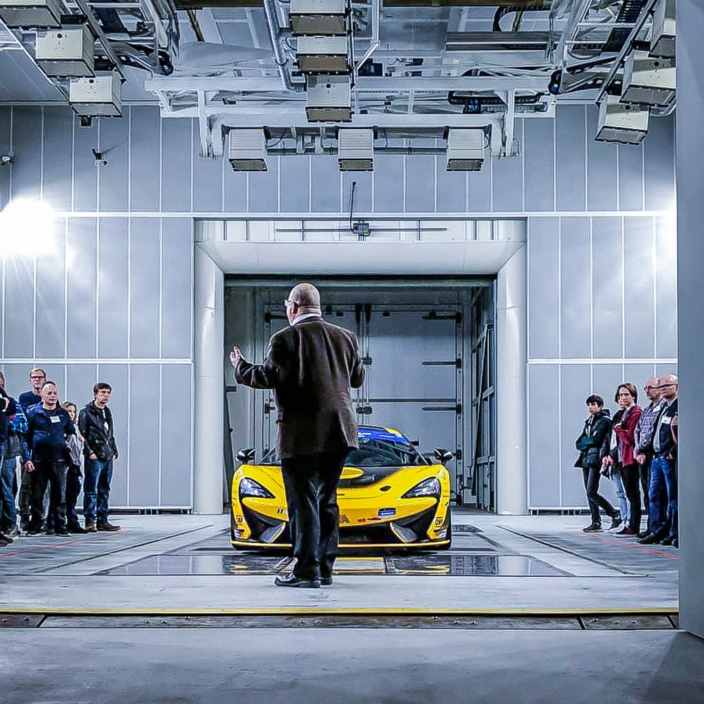 Man stands brightly lit in front of a yellow sports car, speaking to a crowd of individuals to the right and left. 