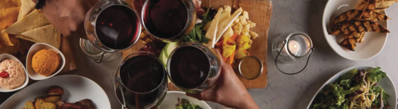 four hands with glasses of wine cheers over breads, salads and various dips and spreads