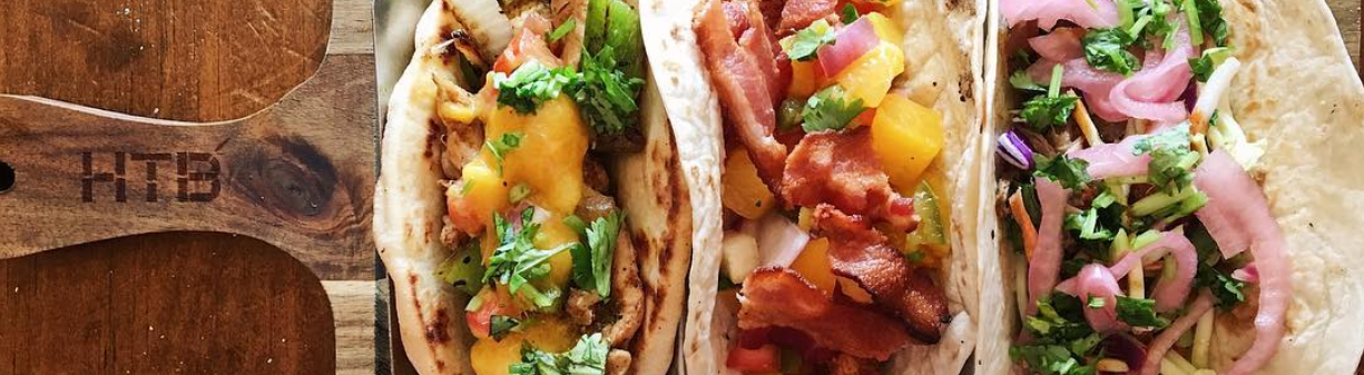 Three tacos line a wooden paddle with the letters HTB engraved on the left side