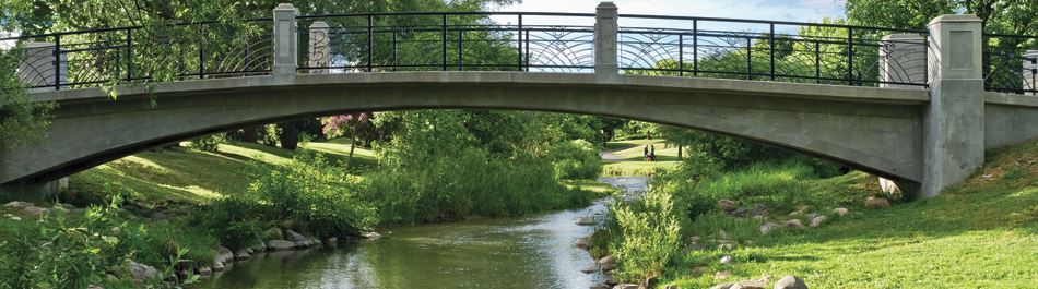 A bridge with black detailing and cement arch crosses over a stream with small and medium size stones with green grass and green trees surrounding.