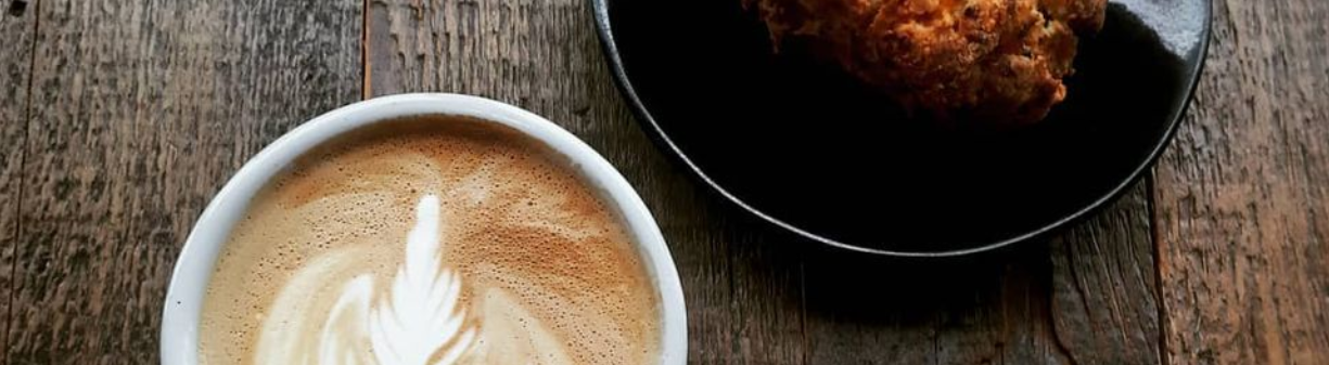 Two cups of coffee fill the screen, both cut off half way. The left cup is frothy with a latte art design, and the right a darker cup of coffee in a black cup with a black saucer. 