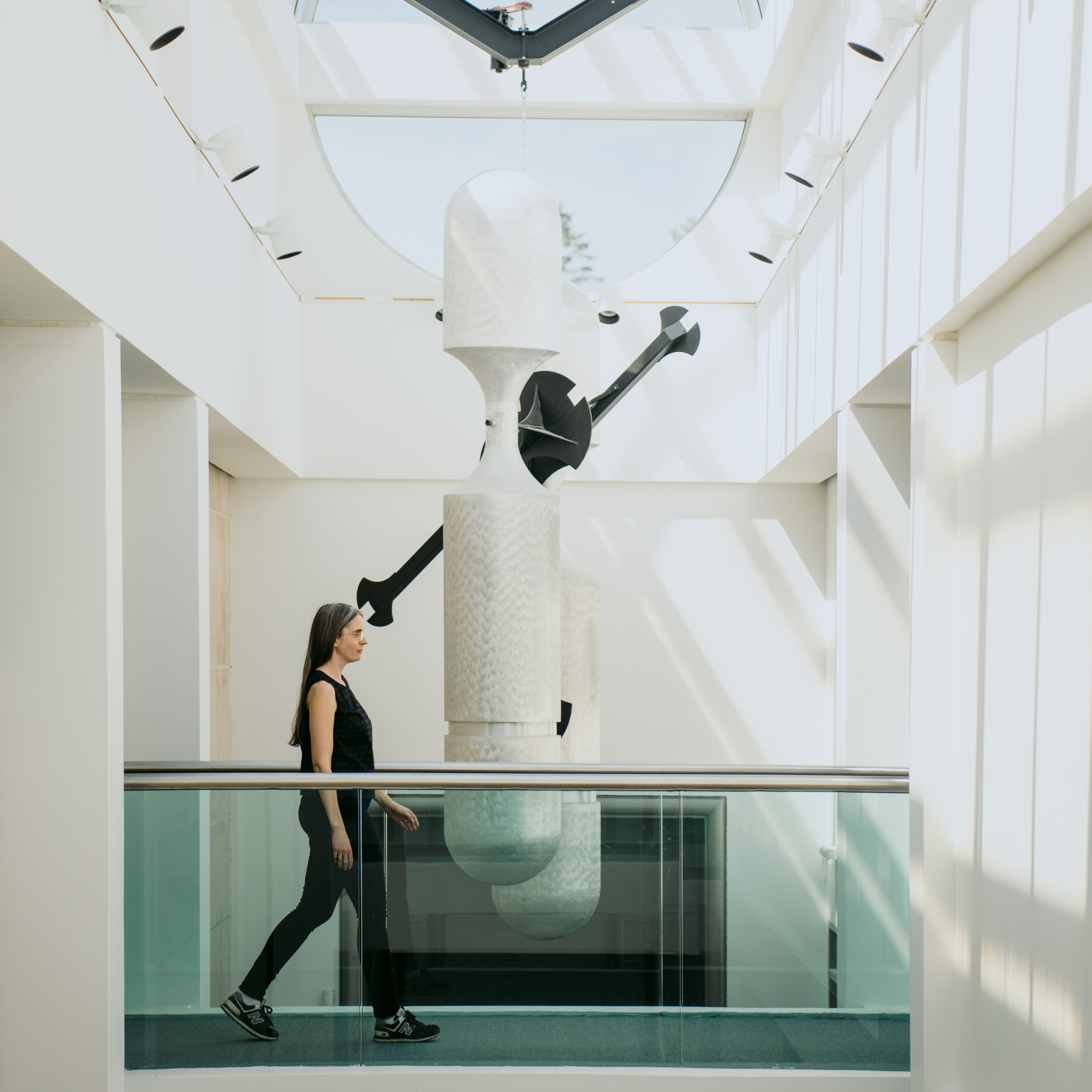 Woman walking along narrow hallway with glass barriers in white building with tall windows from floor to ceiling behind