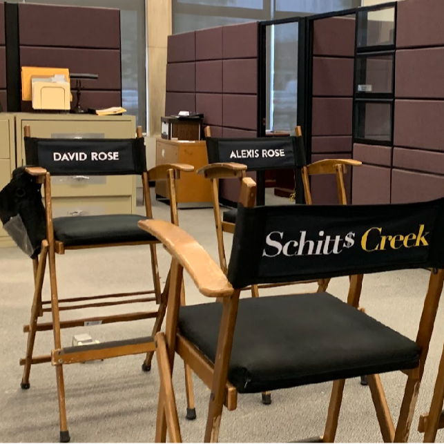 Black film cast chairs are setup surrounding maroon office cubicles and filing cabinets. The back of the chair in the forefront reads "Schitts Creek" and character names on chairs across read character names "David Rose" and to the right "Alexis Rose" 