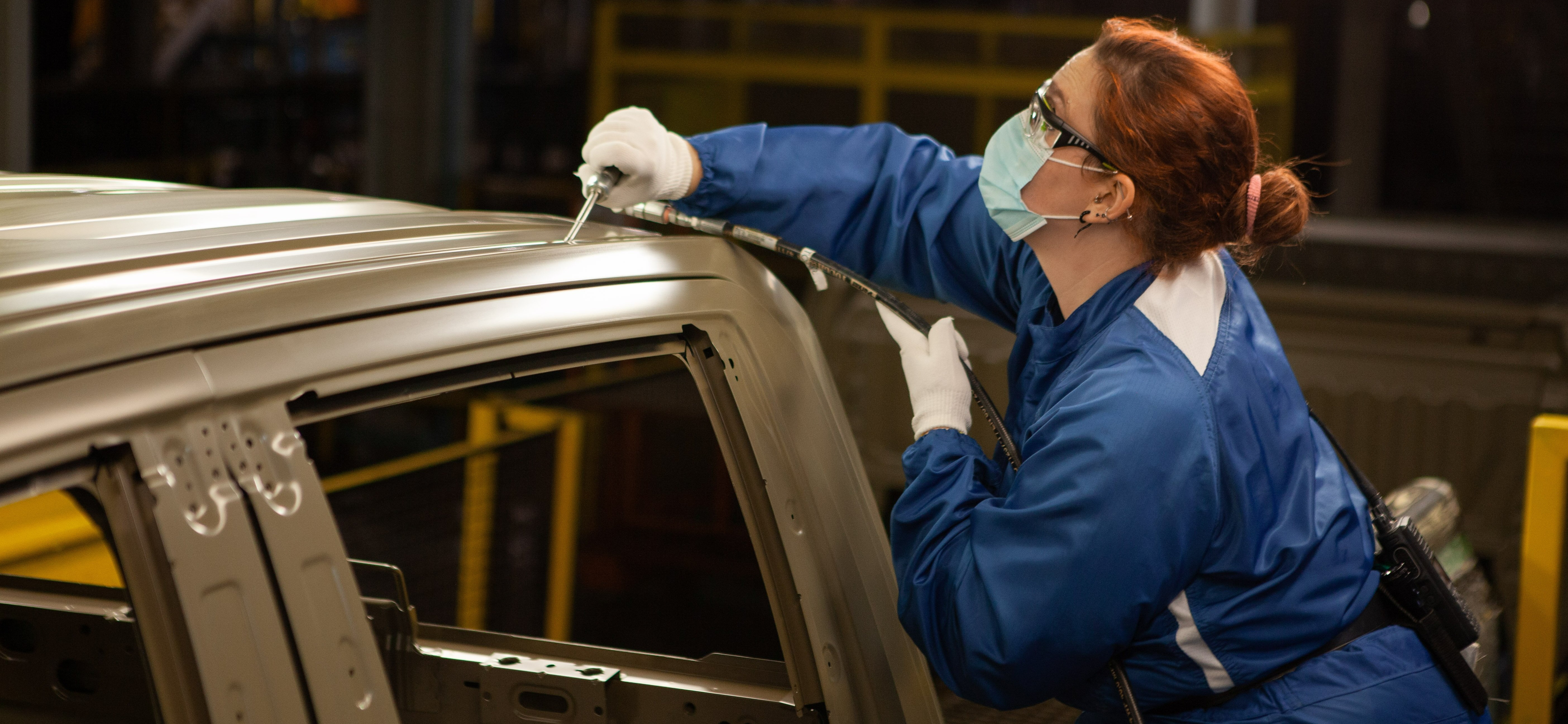 A women wearing a blue facemask uses a hose like piece of equipment on the top of the frame of a vehicle. 