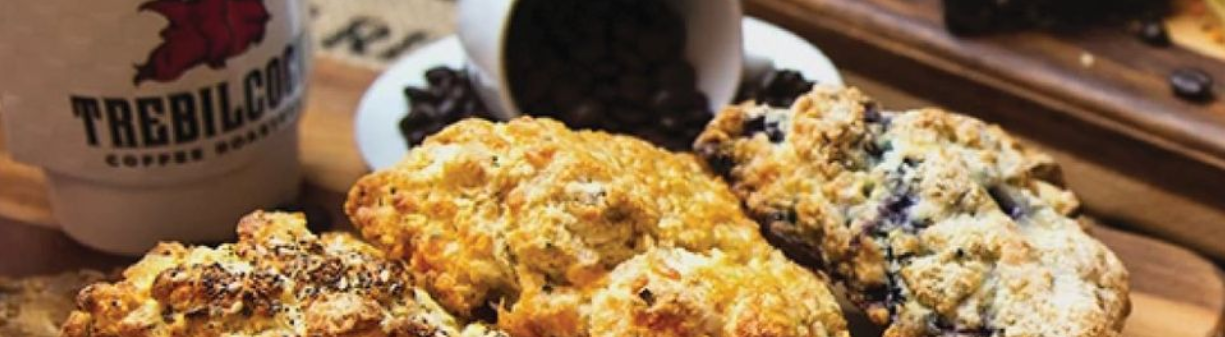 A white coffee cup with the Trebilcock logo on the right, with spilling coffee beans in the background with various types of scones at the foreground. 