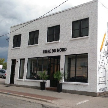 A 2 floor square white building with square windows on the top floor, with black letters reading "Frere Du Nord" with beige sidewalk and two black planters in front 