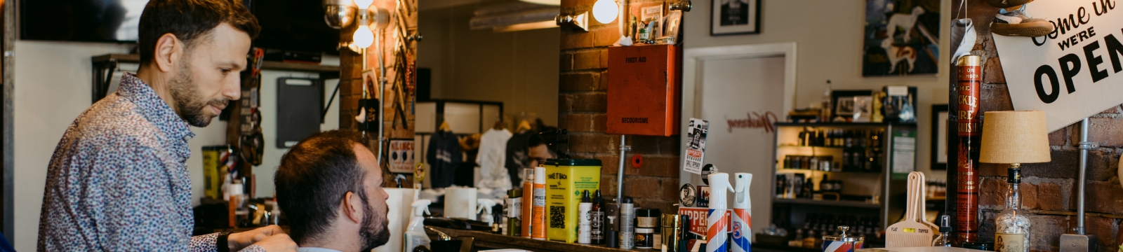 Barber stands behind client in a barbers chair in a barber shop with brick walls and various vintage products surrounding. To the right there is a white sign that reads "come in, we're open."