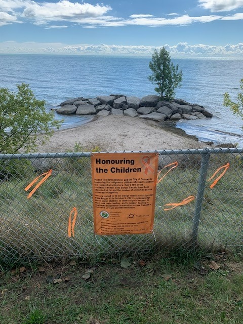 Image of Lakeview Park Orange Ribbon Memorial. View of Lake Ontario and set up on a chain link fence is a banner explained the memorial and orange ribbons that have began to be tied to the chain links 