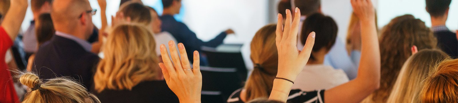 The backs of people in a meeting, some with their hands raised and others without their hands raised.