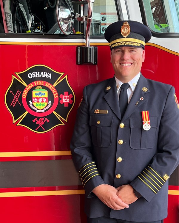 Chief Barkwell smiling looking at the camera while standing in front of an Oshawa Fire Services pumper truck.