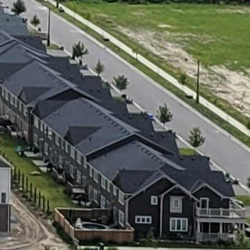 View of brown townhomes with black roofs along a bare road and a grassy field on the right side of the road