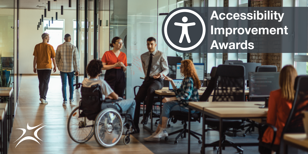 Two people walk away from the camera on the left side of the frame while a group including one person standing, one person leaning, another sitting in a chair and one in a wheelchair sit are having a discussion beside a bank of desks. On the right hand side is the back of a person typing at a computer. The "Accessibility Icon" is in the upper right hand corner with the words: "Accessibility Improvement Awards"