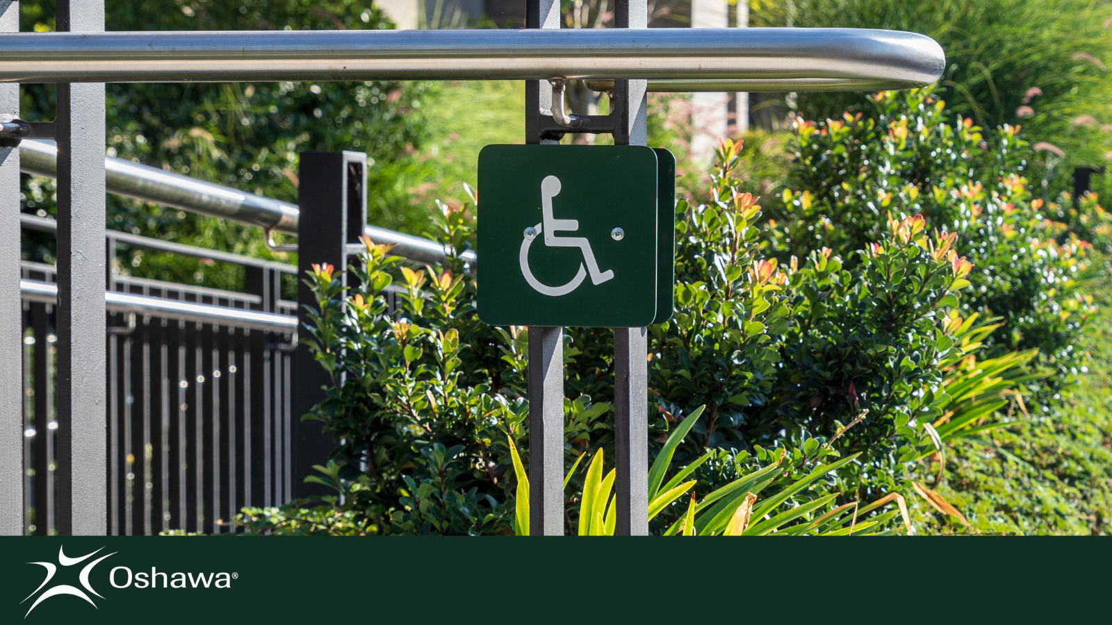 A silver handrail. Below it sits a green accessibility sign. In the background of the photo there is a community garden. The City of Oshawa logo in white appears below on a green background.
