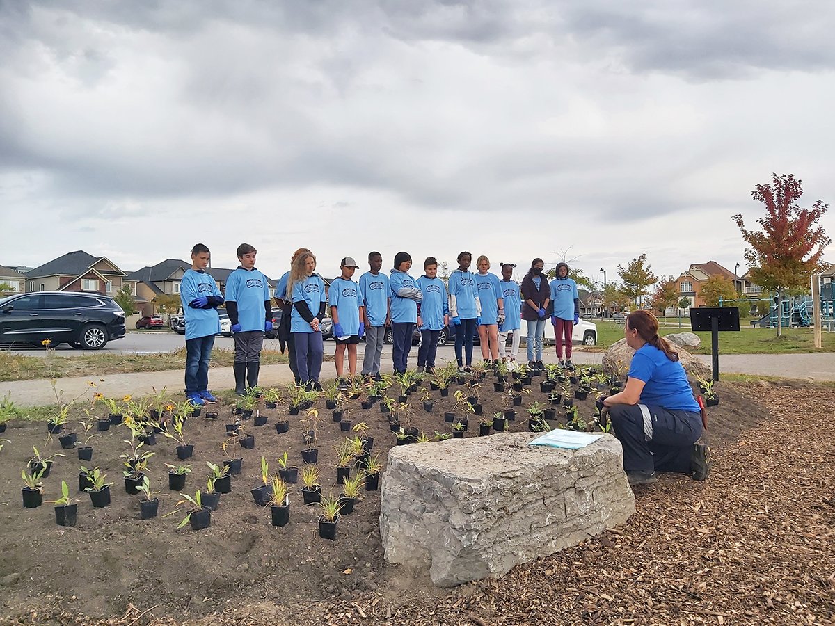 a group of students stand behind a flower bed while a City staff member provides instructions