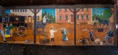 This mural is a streetscape with a black horse drawn carriage, two buildings with one building having "Oshawa House" written on it, and people along a boardwalk with suitcases and parcels.