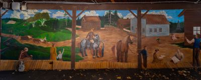This mural is a streetscape depicting the four corners in Oshawa with Skae's checkerboard general store, and two other buildings. People are in front of the buildings along a boardwalk. One person is riding a grey horse.
