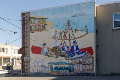 This mural depicts the Lake Ontario waterfront found in Oshawa, before it became Lakeview Park, with a street trolley, people on the beach, and a boat in the background.