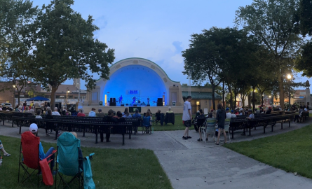 Bandshell stage with concert and individuals sitting watching stage performance.