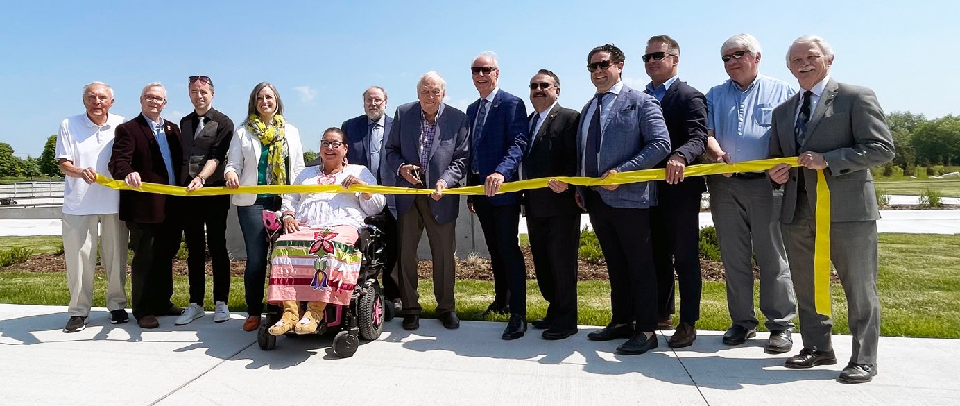 Members of Oshawa City Council and dignitaries with Mr. Ed Broadbent. All are holding onto a yellow ribbon with one hand. Mr. Broadbent is posed holding scissors over the ribbon before cutting it.