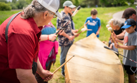 Artist surrounded by children bang on a canoe drum with drumsticks