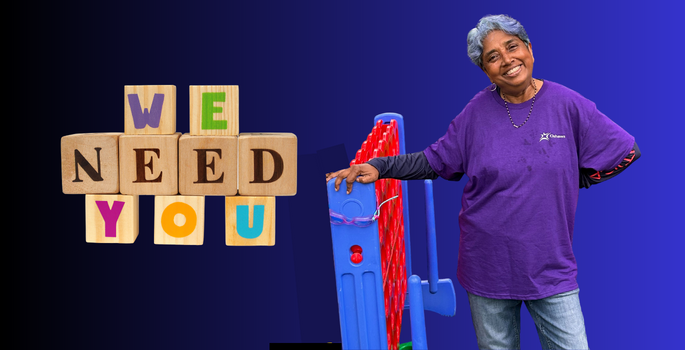 a woman in a purple volunteer shirt standing beside a large plastic game with the words we need you