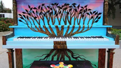 Image of a painted piano and bench. The piano is painted blue with purple and pink clouds along the top. There is a tree growing up from the base of the piano. The tree's branches don't have leaves, instead the branches are full with blackbirds. The piano bench is painted to look like a galaxy and a detailed colour wheel.