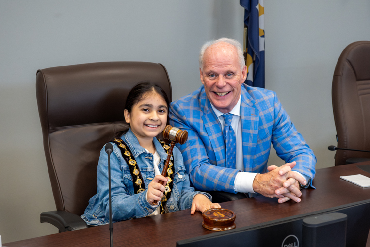 A child holds a gavel while sitting beside the Mayor in City Council chamber office.