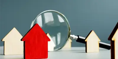 A single red wooden house token stands amongst natural wood house tokens and a magnifying glass