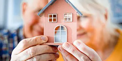 Two seniors hold a wooden house in front of their faces