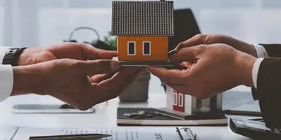 Two people passing a wooden house figurine across a table to one another