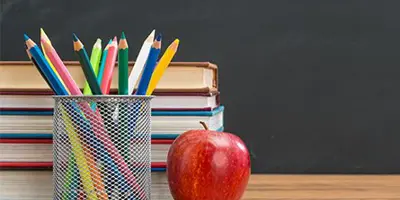 An apple sitting on a desk beside pencil crayons and in front of a stack of books